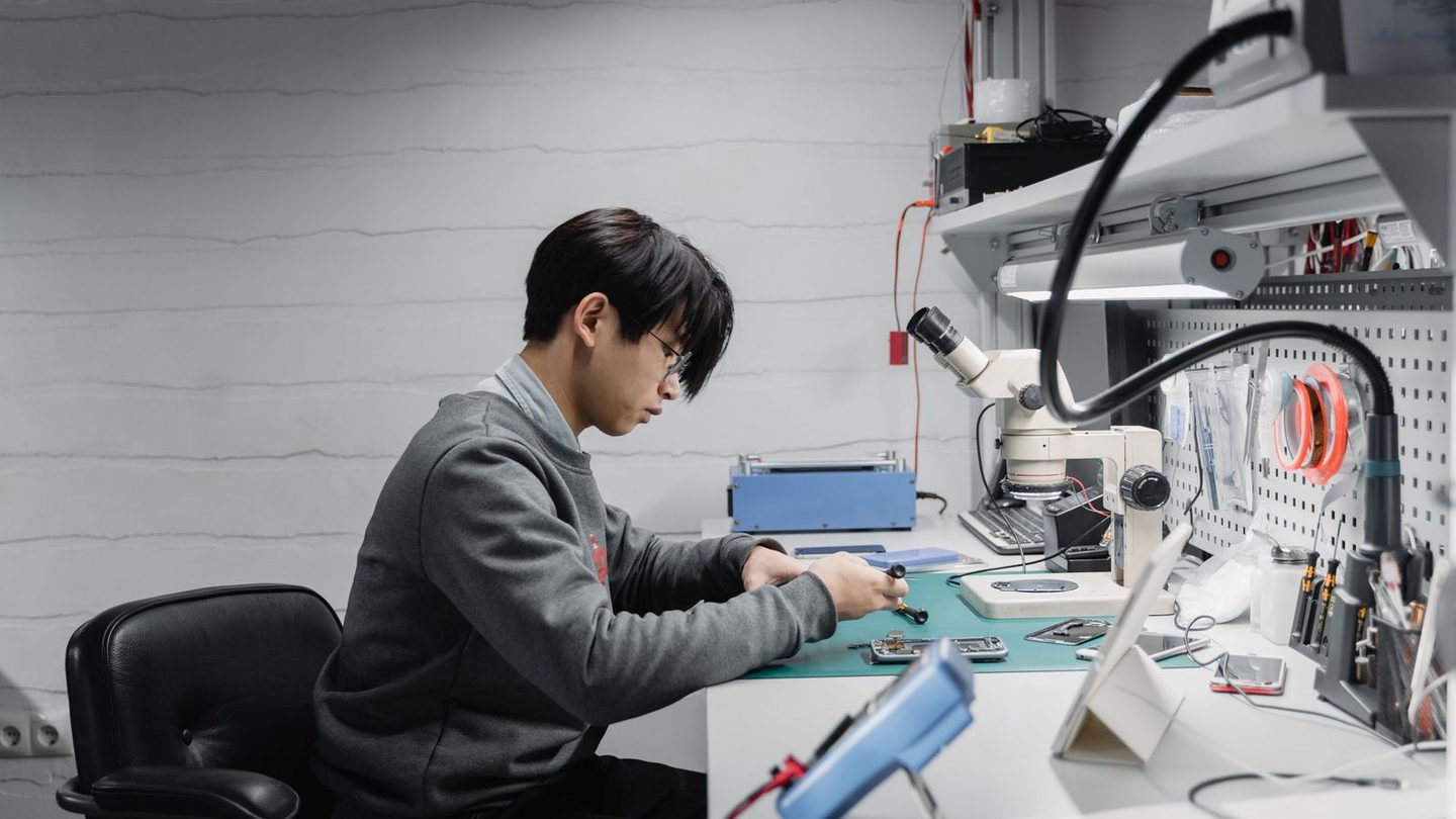An engineer at a bench taking apart a device under a stereo microscope, a multimeter in the foreground